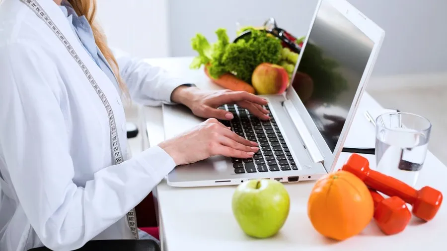 Dietetyczka pracująca przy laptopie, otoczona zdrowymi produktami i sprzętem do ćwiczeń